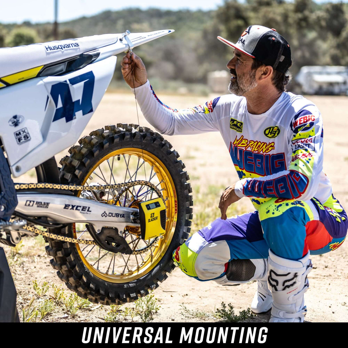 Motocross rider adjusting a Husqvarna bike with slacker suspension device, wearing colourful racing gear.